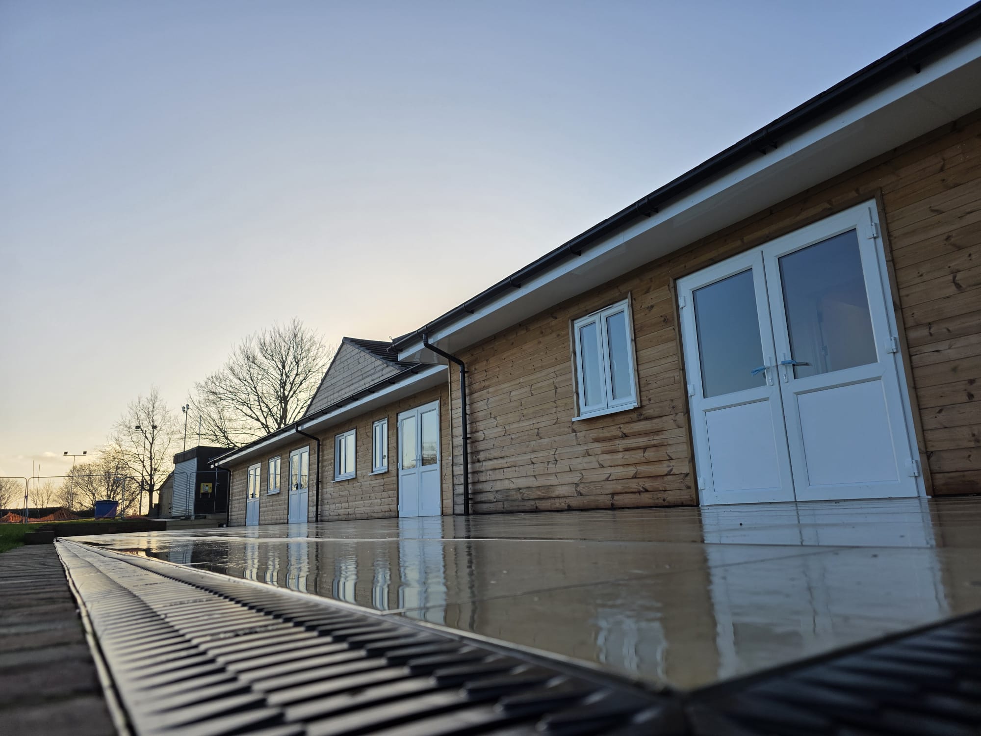 Sports pavilion exterior at dusk showing new timber cladding, double doors and tiled terrace with drainage channel
